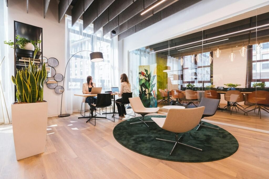 Two people working at laptops by a tall window in a bright modern office, with lounge chairs and a glass meeting room