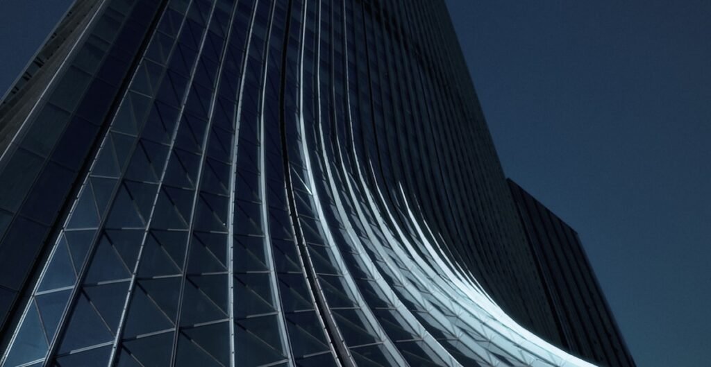 Looking up a dark curved glass skyscraper at night, its ribbed facade catching light beside a second tower
