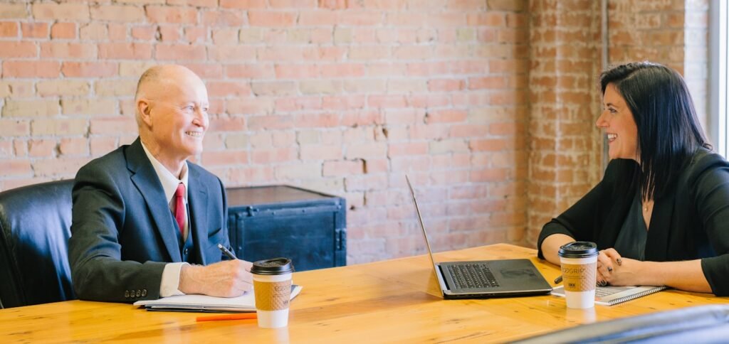 An older man in a suit taking notes across a boardroom table from a woman with a laptop, exposed brick wall behind them