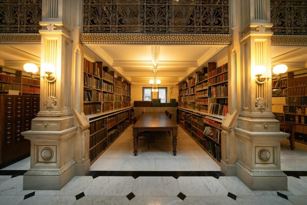 Long wooden reading table between tall shelves of bound volumes in a warmly lit marble library hall