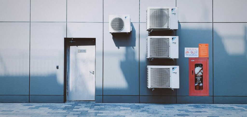 Four white air-conditioning condenser units mounted on the grey panelled wall of a modern commercial building, beside a red fire hose cabinet