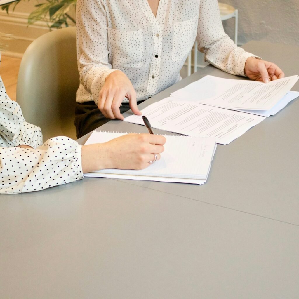 Two people seated at a pale table, one writing with a pen on a notepad beside stacked contract pages