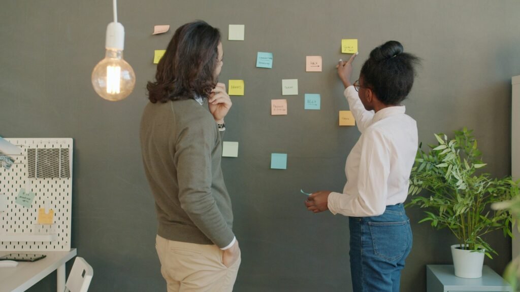 Two colleagues seen from behind reviewing colourful handwritten sticky notes arranged across a grey office wall