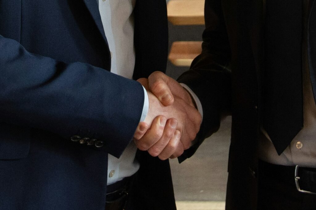 Two people in dark suits shaking hands indoors, close on the clasped hands, warm wooden stairs behind