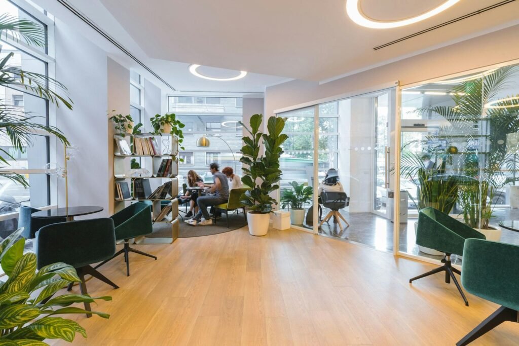 Sunlit office lounge with pale wood floor, green armchairs and palms; three colleagues talk at a table beyond