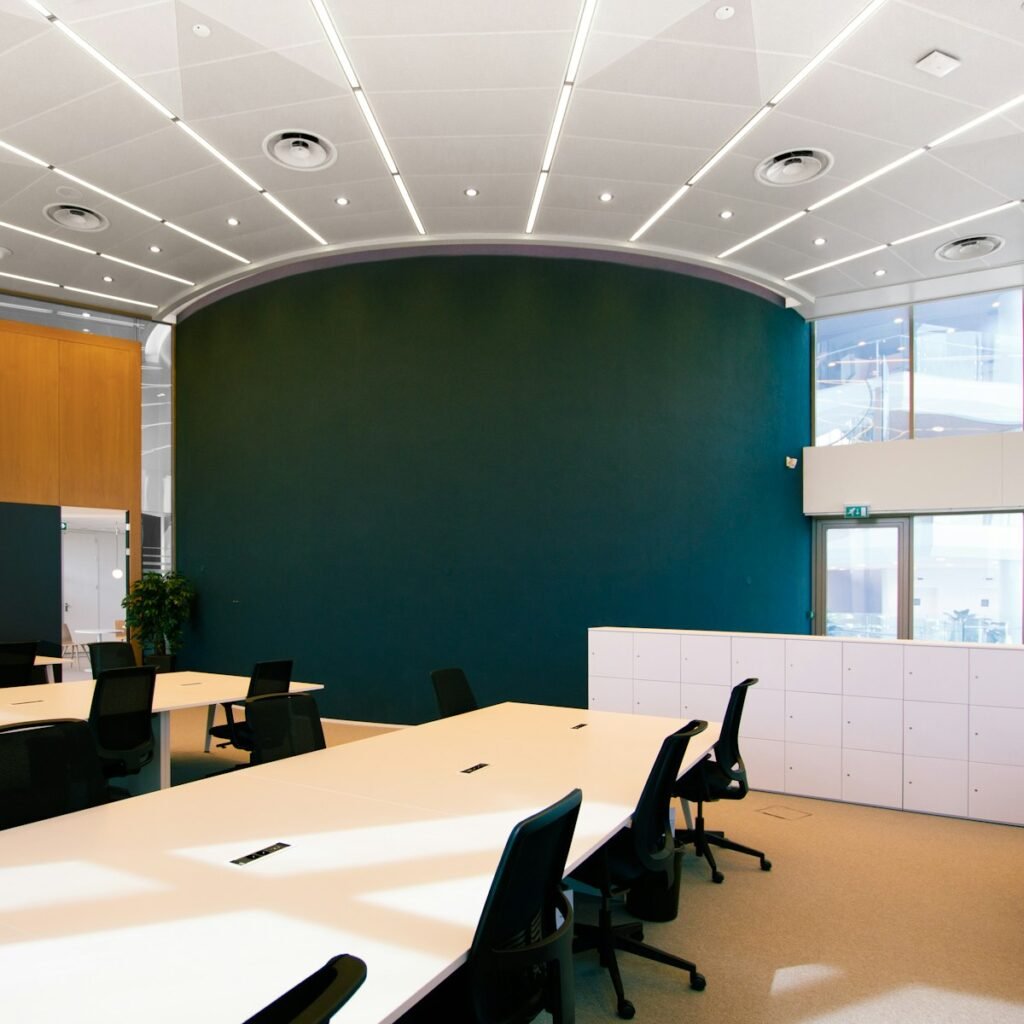 Empty office meeting space with long white tables, black chairs and a curved deep teal wall lit by daylight