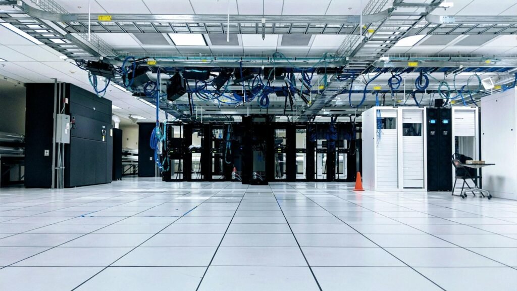 Wide view across a bright data centre floor, dark server racks centred beneath blue cable trays
